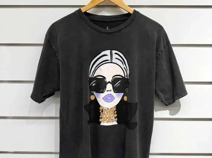 Black graphic tee with stylish woman print, oversized sunglasses, and gold jewelry, hanging on wood hanger