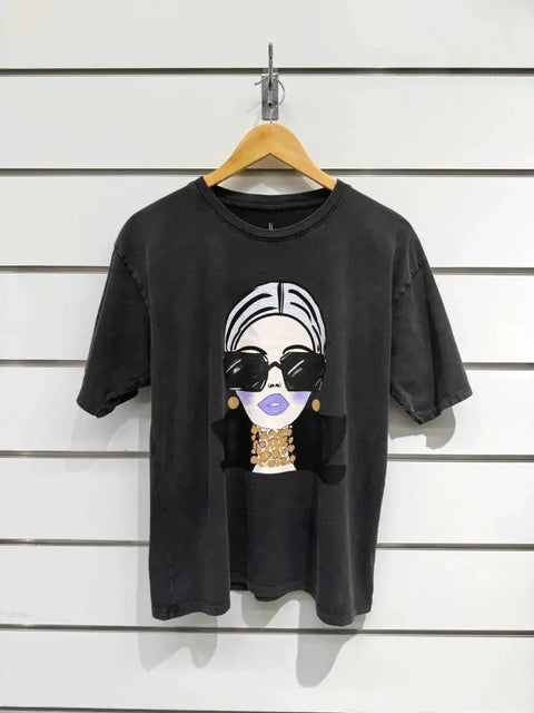 Black graphic tee with stylish woman print, oversized sunglasses, and gold jewelry, hanging on wood hanger